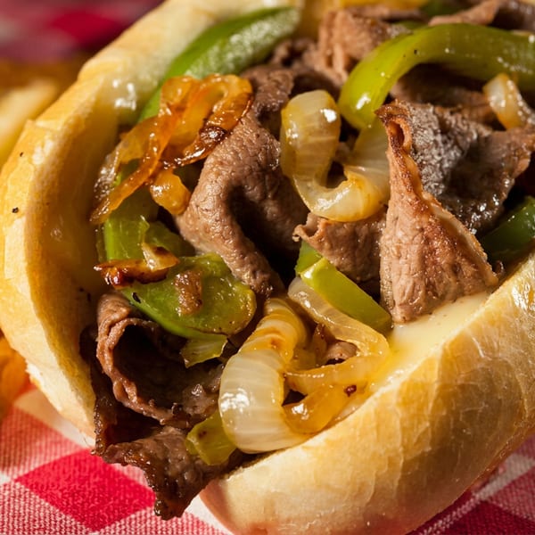 Cheesesteak sandwich with steak, peppers, onions, and melted cheese, served with fries on a red checkered tablecloth.